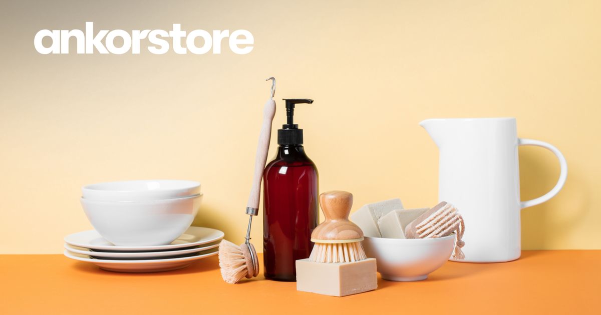 Minimalist still life with Ankorstore logo: stacked white plates and bowls, natural bristle dish brushes, an amber pump bottle, soap bars, and a white pitcher on an orange surface against a yellow backdrop.