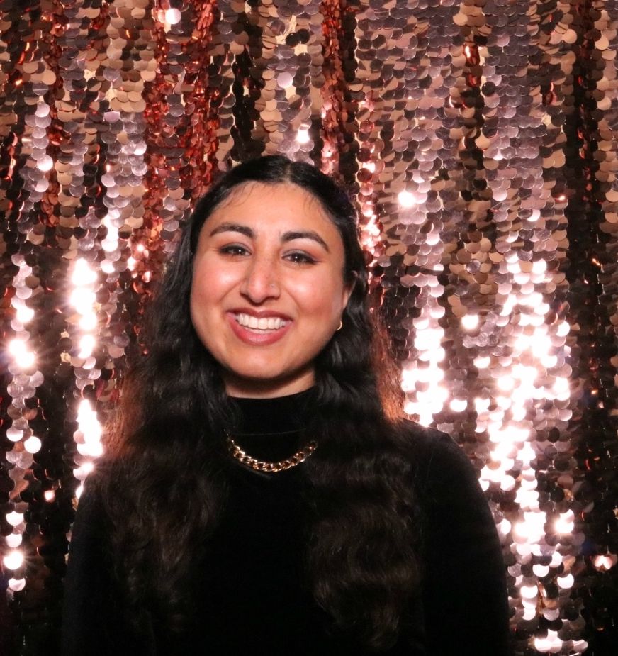 Smiling person with long dark hair in a black top and gold chain necklace, posed before a shimmering rose-gold sequin wall; warm, soft lighting creates bokeh-style sparkles and a festive, polished mood.