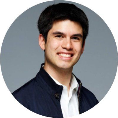 Professional headshot of a smiling man with short dark hair, wearing a navy jacket over a white collared shirt, posed against a neutral gray background in a circular crop.