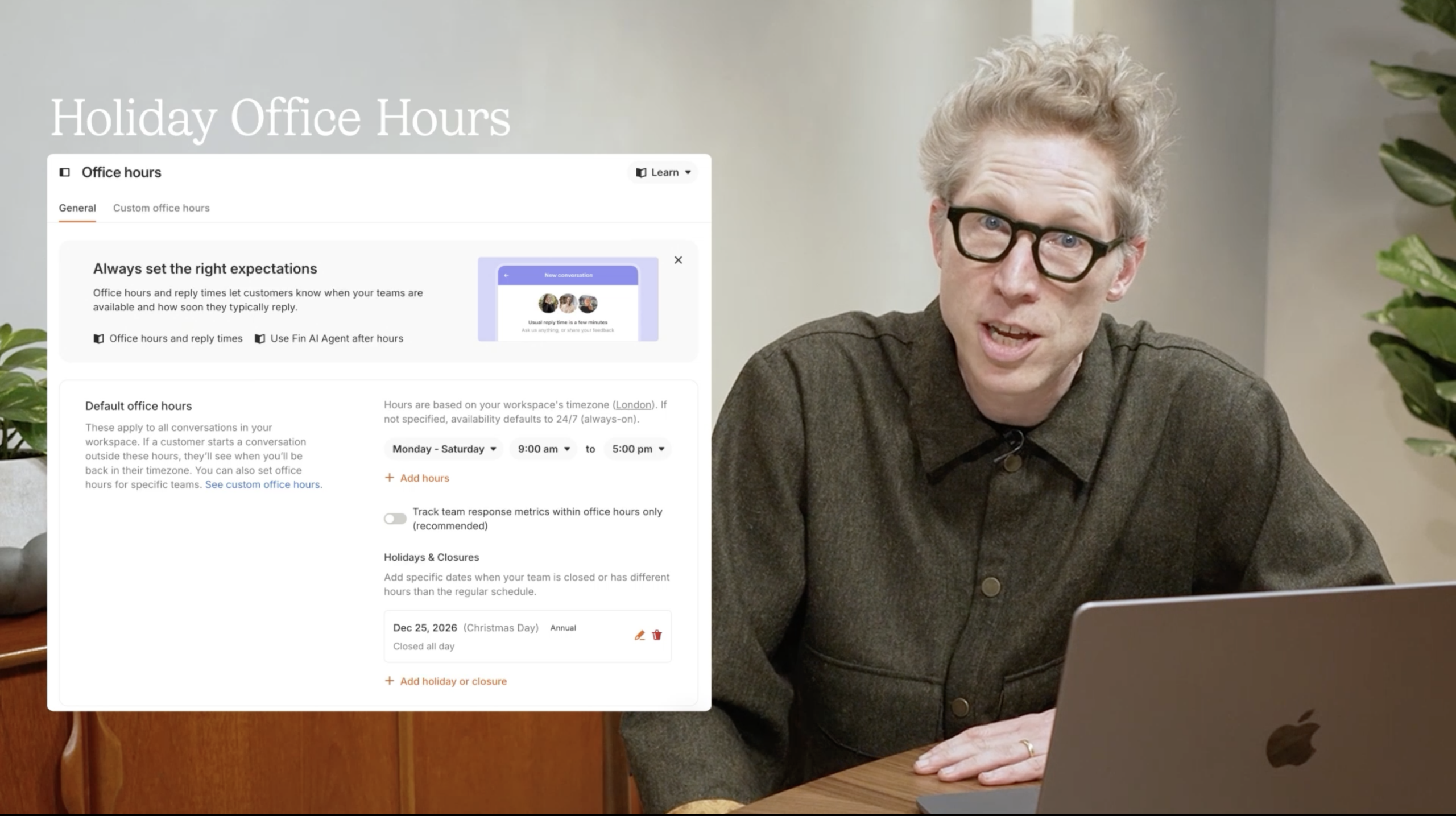 Presenter explains Fin's Holiday Office Hours feature beside a laptop, with a UI screen showing office hours, reply times, and holiday closures settings for customer support teams.