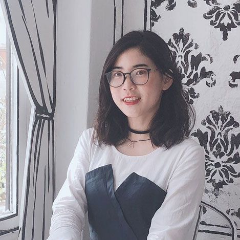 Portrait of a person with shoulder-length dark hair and glasses seated by a bright window, wearing a white top with dark panel and choker, against black-and-white illustrative wallpaper.