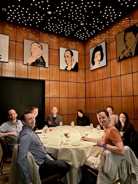 Group of colleagues seated around a round table in a warm wood-paneled private dining room with pop-art portraits and a starry ceiling, smiling over drinks and dessert as they mark a business milestone.